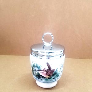 May include: White ceramic egg cup with a silver lid and a small bird illustration. The bird is brown, white, and green and is perched on a branch with green leaves.