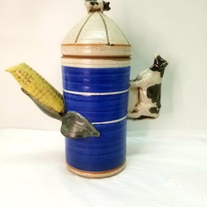 May include: A blue and white ceramic pitcher with a cow on the side and a corn cob spout. The pitcher has a white lid with a small cow on top.