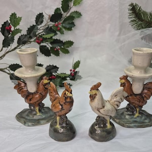 May include: Four ceramic rooster candle holders. Two have a candle cup and saucer, and two are standalone rooster figurines. The roosters are brown, white, and tan, with a green and brown base. Decorative holly and greenery are in the background.