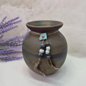 May include: A round, dark-toned ceramic vase with a matte finish, featuring a decorative cord tied around the neck. The cord is adorned with a turquoise-colored stone, various blue and white beads, and two brown-toned feathers hanging down.