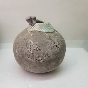 May include: A gray ceramic vase with a crackled glaze and a light green and purple rim. The vase has a rounded shape and a small opening at the top.