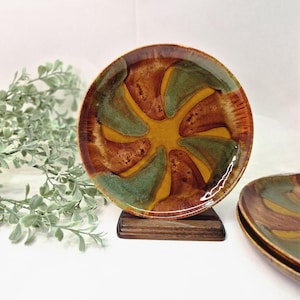 May include: A ceramic plate with a unique design. The plate features a spiral pattern with shades of brown, green, and yellow. The plate is displayed on a wooden stand, showcasing its artistic design. The plate is a decorative piece.