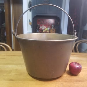 May include: A vintage copper bucket with a metal handle. The bucket is brown and has a worn, rustic look. It is sitting on a wooden surface.