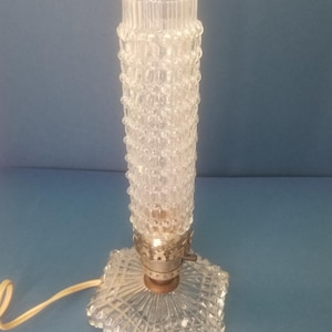 May include: Clear glass table lamp with a textured, cylindrical shade and a square base. The lamp has a decorative, ridged top and a metal base. The lamp is plugged in and has a yellow cord.