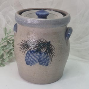 May include: A cylindrical stoneware crock with a speckled beige finish, featuring a blue painted pinecone and needle motif on the front. The vessel includes two side handles, a matching lid with a blue knob, and a blue rim around the top edge.