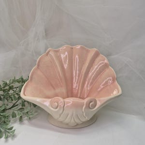 May include: A pink ceramic seashell-shaped decorative bowl. The bowl has a scalloped edge and a cream-colored base. The interior of the bowl is a soft pink color. The bowl is designed to be a decorative piece.