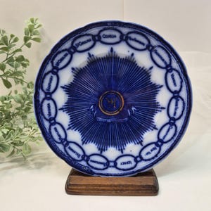 May include: A vintage blue and white decorative plate with a detailed design. The plate features a central starburst pattern and is encircled by the names of U.S. states. The plate is displayed on a small wooden stand.