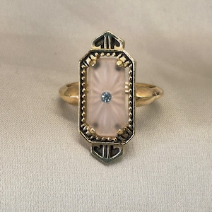 May include: A gold ring featuring an elongated octagonal setting with a frosted pink glass center. The glass is accented by a small, central light blue stone. The metal frame has a dark, recessed finish with beaded edges and pointed ends.