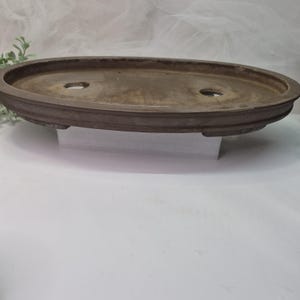 May include: A brown, oval-shaped bonsai pot with two drainage holes. The pot has a raised, decorative rim and is made of a weathered material. The pot is designed for planting and displaying bonsai trees.
