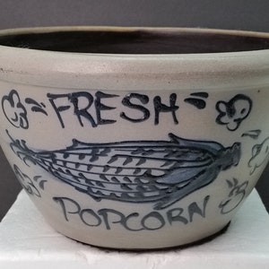 May include: A white ceramic bowl with a blue and black hand-painted design of an ear of corn and the words "FRESH POPCORN".