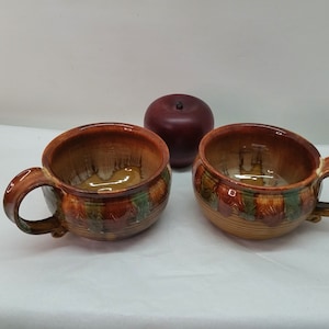 May include: Two ceramic soup bowls with handles, featuring a brown and tan color scheme with green and red accents. A dark red decorative apple sits between the bowls. The bowls have a rustic, handcrafted appearance.