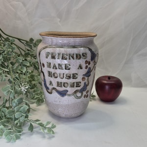 May include: A speckled ceramic vase with the words "FRIENDS MAKE A HOUSE A HOME" in dark brown lettering. The vase has a light brown rim and is decorated with blue and green floral accents. A red apple sits beside the vase.