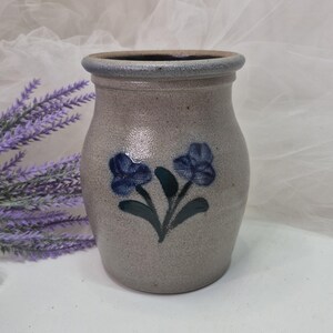 May include: A stoneware crock with a speckled light gray finish and a smooth, rounded rim. The front features a hand-painted design of two dark blue flowers with green stems and leaves. The interior is dark, and the exterior has a textured surface.