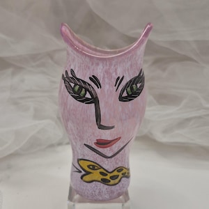 May include: A pink and white speckled art glass vase with a face design. The vase features a stylized face with black eyelashes, green eyes, a black nose, and red lips. A yellow and black bow tie is at the base.