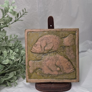 May include: A square, ceramic tile featuring a relief of two fish. The fish are a salmon color against a green background. The tile is displayed on a wooden stand. The overall aesthetic is rustic and earthy.
