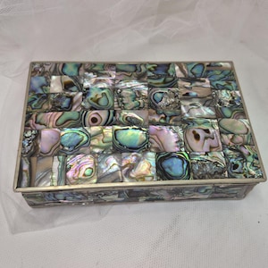 May include: A rectangular box featuring a mosaic of iridescent abalone shell tiles in shades of blue, green, and purple. The lid and sides are covered in these shimmering, multi-colored shell pieces, framed by a thin, metallic silver-toned border.