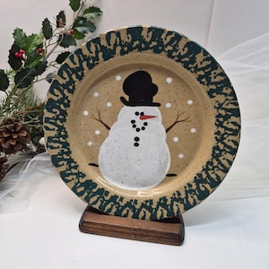 May include: A ceramic plate featuring a snowman design. The snowman has a black top hat, a carrot nose, and coal eyes and buttons. The plate has a speckled tan center and a dark green patterned border. The plate is displayed on a wooden stand.
