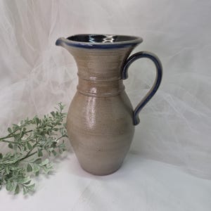 May include: A handmade stoneware pitcher with a tan body and a dark blue rim and handle. The pitcher has a classic shape with a spout for easy pouring. The handle is curved and comfortable to hold. A decorative piece for the kitchen or dining room.