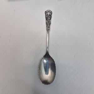 May include: A silver spoon with an ornate handle and a scalloped bowl. The handle features detailed scrollwork and a decorative finial. The spoon is likely made of sterling silver and is designed for serving or eating.
