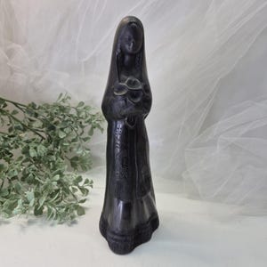 May include: A dark, carved statue of a woman wearing a veil and holding a bouquet of flowers. The statue has intricate details on the clothing and is set against a soft, white background.