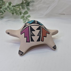 May include: A tan-colored ceramic turtle figurine with a painted design. The turtle's shell features a geometric pattern in black, purple, orange, and turquoise. The turtle has four legs and a head, all extending from the body.