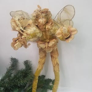 May include: A whimsical fairy figurine with a smiling face, adorned in shades of gold and tan. The fairy has large, delicate wings and is holding flower-like embellishments. The figure is standing, with long, slender legs, and is decorated with leaf-like details.