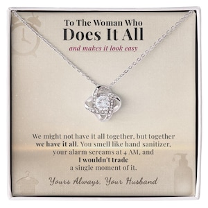 Könnte beinhalten: Eine silberfarbene Halskette mit einem knotenförmigen Anhänger, besetzt mit klaren Kristallen. Die Halskette wird auf einer Karte mit dem Text "To The Woman Who Does It All" präsentiert. Die Karte enthält auch eine liebevolle Nachricht von "Your Husband".