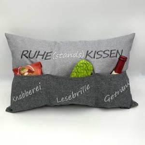 Könnte beinhalten: Ein graues Kissen mit drei Taschen und dem aufgenähten Text "RUHE (stands) KISSEN" auf der Vorderseite. Die Taschen sind mit "Knabberei", "Lesebrille" und "Getrank" beschriftet.
