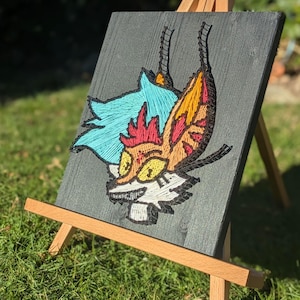 May include: A colorful string art piece depicting a stylized fox head with blue, orange, yellow, and red accents. The artwork is mounted on a dark gray wooden board and displayed on a small wooden easel, set against a backdrop of green grass and foliage.