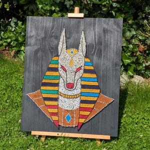 May include: A string art piece depicting Anubis, the Egyptian god of the afterlife, against a black wooden background. The artwork features a detailed design with colorful thread, including red, yellow, blue, and orange, mounted on a wooden easel.