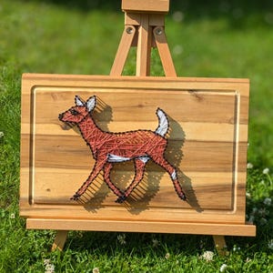 May include: A wooden cutting board with a deer string art design. The deer is crafted with red string and features white accents on its ears and tail. The board is displayed on a small wooden easel, set against a backdrop of green grass.