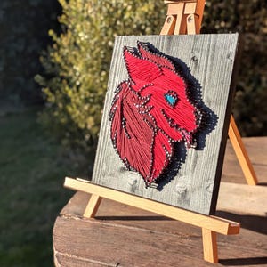 May include: A red string art piece depicting a wolf's head with a teal eye, mounted on a gray wooden board. The artwork is displayed on a small wooden easel. The piece is set against a blurred outdoor background.