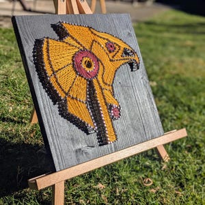 May include: A string art piece depicting a stylized bird of prey, likely a hawk or falcon, in shades of yellow, black, and burgundy. The artwork is mounted on a gray wooden board and displayed on a small wooden easel, set against a backdrop of green grass.