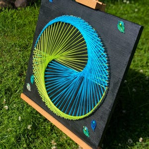 May include: A square, black wooden art piece with a geometric design made of blue and yellow string. The string art is in a yin-yang style, with peacock feather accents. The artwork is displayed on a wooden easel.