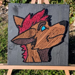 May include: A string art piece featuring a stylized wolf head profile. The artwork uses brown, red, black, and blue thread on a dark gray wooden board. The piece is displayed on a small wooden easel, set against a backdrop of green foliage.