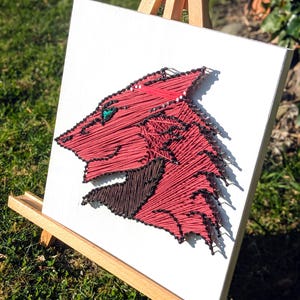May include: A red and brown string art piece depicting a stylized animal head with a green gem eye. The artwork is mounted on a white canvas and displayed on a wooden easel, set against a backdrop of green grass.