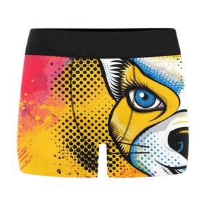 Puede incluir: Calzoncillos tipo bóxer con un gráfico pop art de una cara de gato con un ojo azul brillante, pelaje amarillo y bigotes negros. El diseño incorpora una cinturilla negra, un fondo con salpicaduras amarillas y naranjas y puntos de semitono.