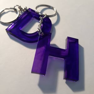 Può includere: Portachiavi in ​​acrilico viola con le lettere "D" e "H" attaccate a una catena e un anello argentati.
