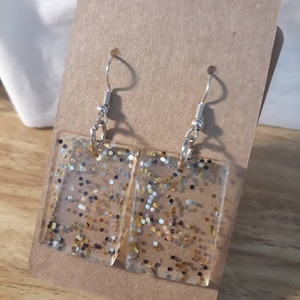 May include: A pair of rectangular dangle earrings with gold, silver, and black glitter embedded in clear resin. The earrings have silver hooks.