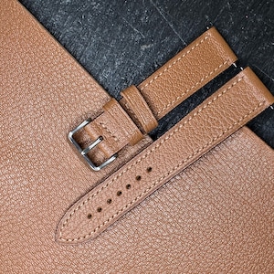 May include: A brown leather watch strap with a silver buckle, lying on a textured brown surface. The strap has a stitched design along the edges and several holes for adjustment. The background is a dark, textured surface.