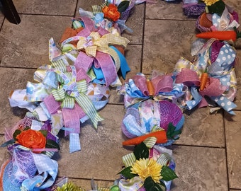 May include: A colorful Easter wreath made of pink, blue, and green ribbon, mesh, and flowers. The wreath is decorated with carrots, eggs, and other Easter-themed decorations.