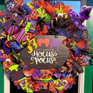 May include: A Halloween wreath with black, orange, purple, and green ribbon and mesh. It features a sign that says "It's all a bunch of Hocus Pocus" and a witch's hat with a black and purple bow.