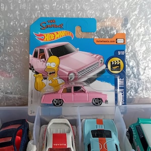 May include: A pink Hot Wheels die-cast car from The Simpsons series, featuring Homer Simpson. The packaging includes the text "The Simpsons" and "Screen Time". The car is 3 inches long.