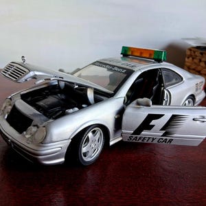 May include: A silver Mercedes-Benz F1 Safety Car model vehicle with an open hood and driver-side door. The car features a roof-mounted light bar, black F1 SAFETY CAR lettering on the door, and Mercedes-Benz branding on the windshield.