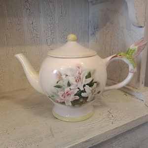 Può includere: Teiera in ceramica bianca con manico a forma di colibrì e design floreale. La teiera presenta uno sfondo bianco con gigli rosa e bianchi e foglie verdi. Il colibrì è verde con accenti rosa.