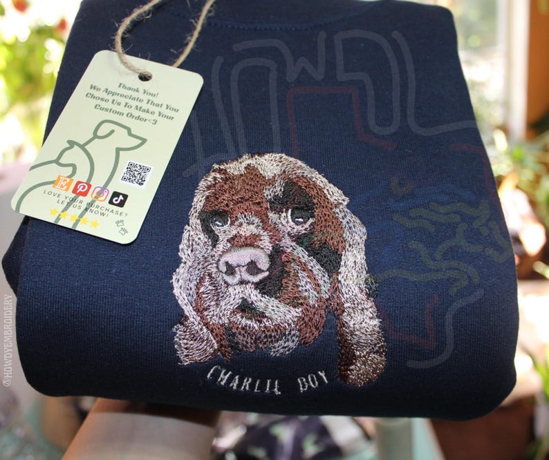 Puede incluir: Una sudadera azul marino con una imagen bordada de un perro marr&oacute;n y blanco. El nombre del perro, "CHARLIE BOY", est&aacute; bordado debajo de la imagen.