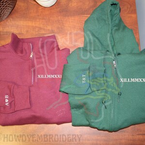 May include: Two sweatshirts, one maroon and one green, with embroidered designs. The maroon sweatshirt has a zipper and the text "A&B" on the sleeve. The green sweatshirt has a zipper and the text "T&T" on the sleeve. Both sweatshirts have the text "XII.I.MMXXIII" on the front.