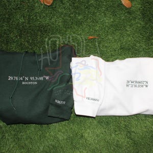 May include: Two sweatshirts with different designs. The green sweatshirt has the text "29.7604 N 95.3698 W HOUSTON EST 205 MMXV" printed on it. The white sweatshirt has the text "29&deg;44'59.6652"N 95*21'30.3156"W VILLMMXVI" printed on it.