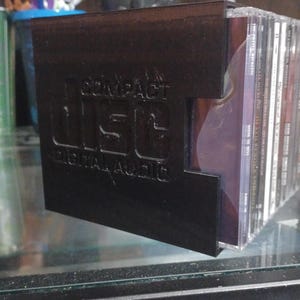 May include: A black plastic storage rack for compact discs featuring the embossed text COMPACT disc DIGITAL AUDIO on the side panel. The rack holds a row of multiple standard jewel cases, allowing the spines of the discs to be visible for identification.