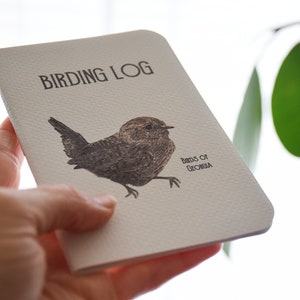 Könnte beinhalten: Ein weißes Notizbuch mit dem Text "Birding Log" und einer Zeichnung eines braunen Vogels mit dem Text "Birds of Georgia".